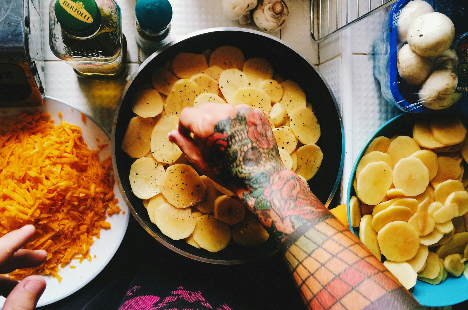 A cook seasoning sliced potatoes arranged in pans beside grated cheese and mushrooms