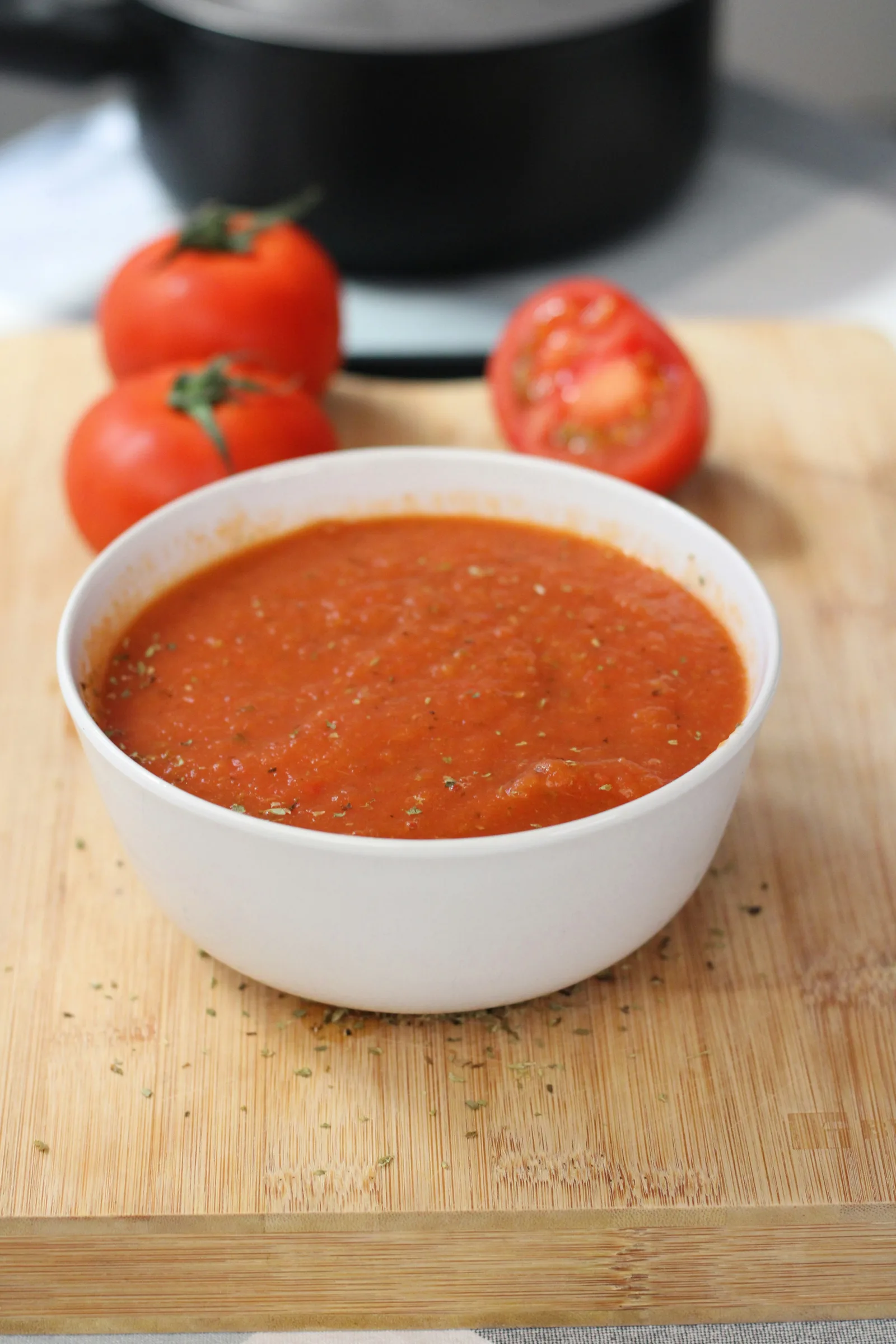 A white bowl filled with smooth tomato sauce beside whole and halved tomatoes